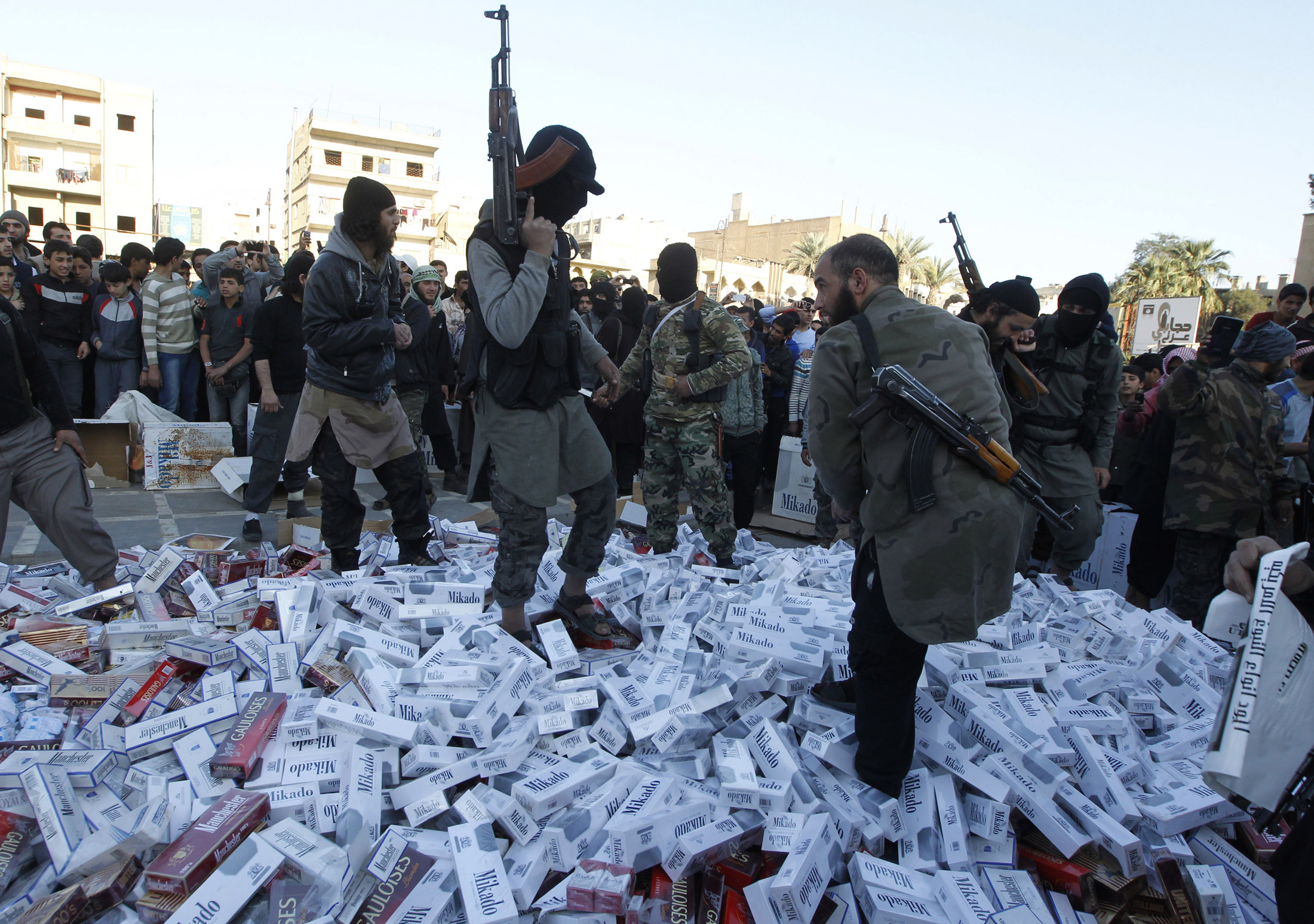 مقاتلون من تنظيم الدولة الإسلامية يحملون أسلحتهم ويقفون على عبوات سجائر مصادرة قبل حرقها، الرقة، سوريا، 2 أبريل 2014. رويترز، صحفي محلي.